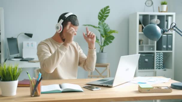 Wesoły pracownik biurowy noszący słuchawki poruszające ramionami tańczące i korzystające z laptopa — Wideo stockowe