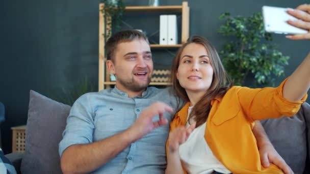 Ευτυχισμένο νεαρό ζευγάρι που παίρνει selfie με smartphone στο σπίτι gesturing πόζα — Αρχείο Βίντεο