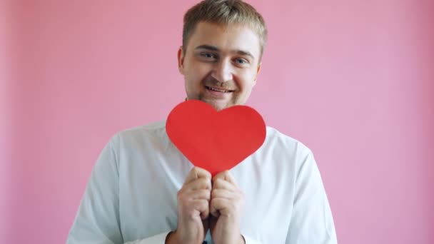 Glimlachende man verstopt gezicht achter hartvorm valentijnskaart draagt elegante kleding — Stockvideo