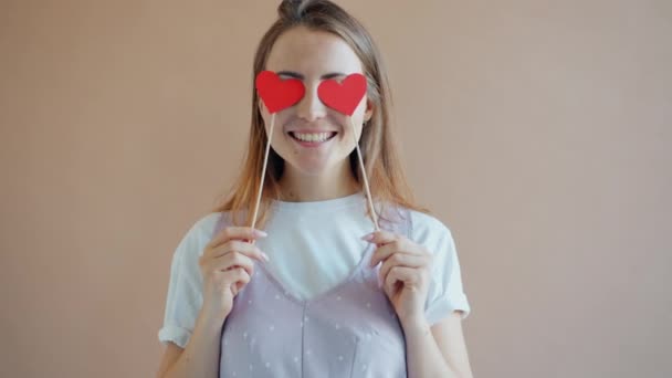 Retrato de una joven alegre sosteniendo formas de corazón ocultando ojos sonriendo — Vídeo de stock