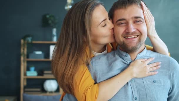 Portrait of happy young woman kissing and hugging husband at home looking at camera — Stock Video