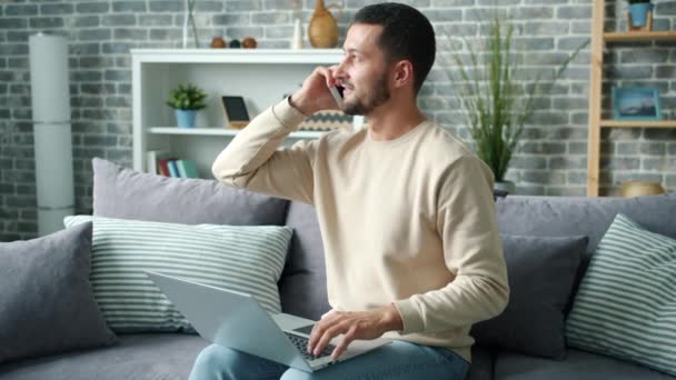 Feliz freelancer hablando por teléfono móvil usando el portátil en el sofá en el apartamento — Vídeo de stock