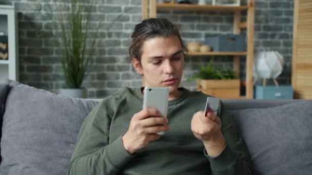 Радостный молодой человек делает покупки онлайн с помощью кредитной карты и смартфона улыбаясь — стоковое видео