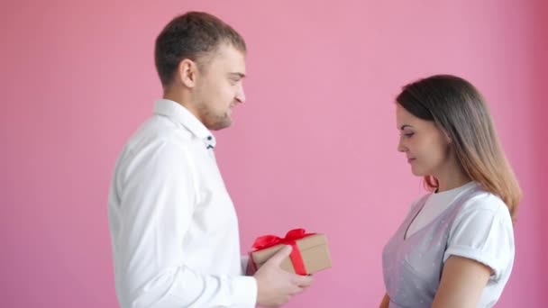 Повільний рух хлопця, що дарує подарункову коробку гарній дівчині, яка цілується на рожевому фоні — стокове відео