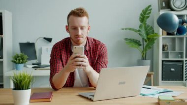 Modern akıllı telefonla iletişim kuran çekici bir adam ofiste tek başına gülümsüyor.