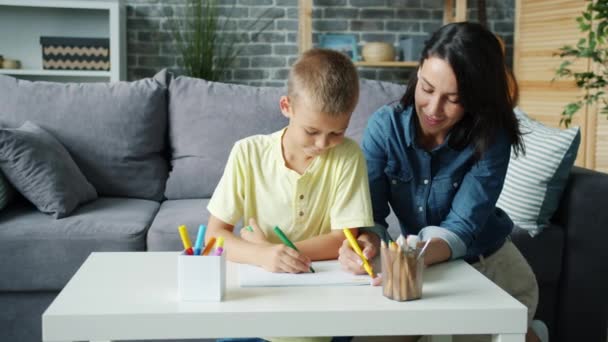 Kleine jongen tekening met stiften samen met zorgzame moeder in gezellige kamer thuis — Stockvideo