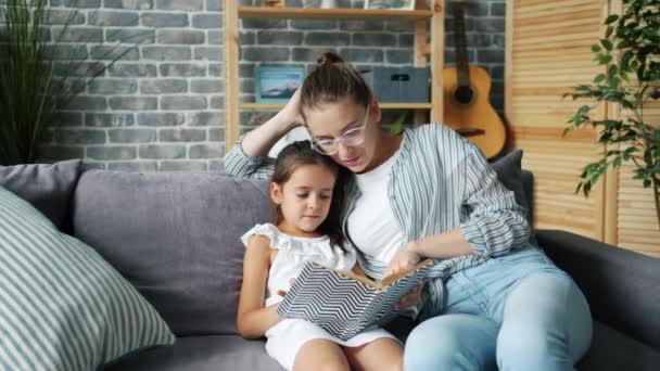 Μητέρα και κόρη διαβάζοντας βιβλίο στο σπίτι χαμογελώντας χαλαρώνοντας στον καναπέ μαζί — Αρχείο Βίντεο