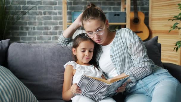 Заботливая мама читает книгу вслух сидя дома на диване с очаровательной дочерью — стоковое видео