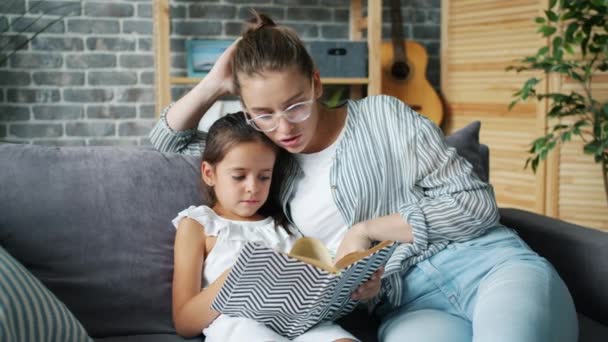 Щаслива молода сім'я мати і дочка читають дитячу книгу вдома посміхаючись — стокове відео