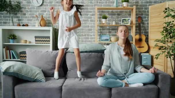 Glad liten flicka hoppar på soffan medan mamma kopplar av i Lotus position hemma — Stockvideo