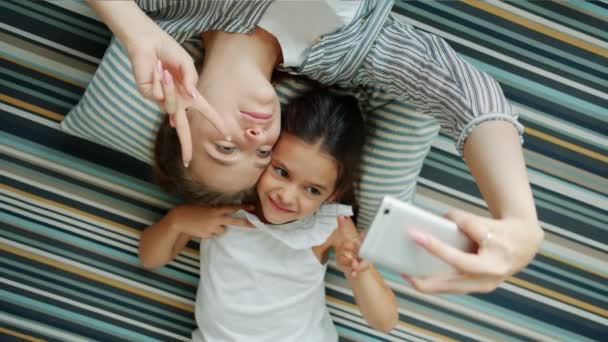 Chica y madre tomando selfie con teléfono inteligente tumbado en el suelo haciendo caras divertidas — Vídeo de stock