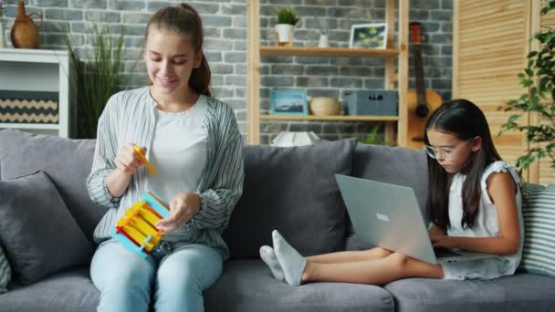 Divertente donna adulta che gioca con il giocattolo mentre figlia seria in occhiali usando il computer portatile — Video Stock