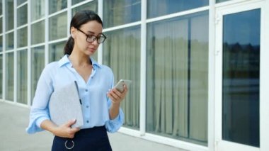 Cam duvar ofis binasının yanında akıllı telefon kullanan kendine güvenen bir iş kadını.