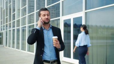 Neşeli girişimci elinde kahve tutarak şehirde yürüyüp cep telefonuyla konuşuyor.