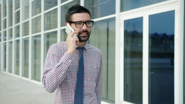 Молодий бізнесмен, що виходить за межі офісної будівлі, розмовляє на мобільному телефоні, посміхаючись — стокове відео