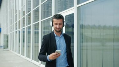 Kulaklıklı rahat bir işadamı dışarıda yürürken akıllı telefon gülümsüyor.