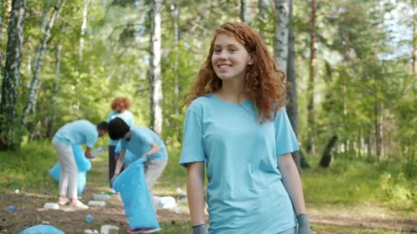 Портрет молодой эко-активистки, улыбающейся в лесу, пока команда собирает мусор — стоковое видео