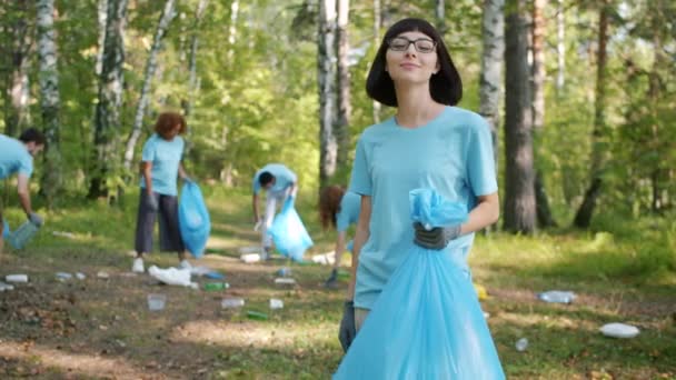Menina sorridente bonito com saco de lixo eco voluntário transformando-se para câmera na floresta — Vídeo de Stock