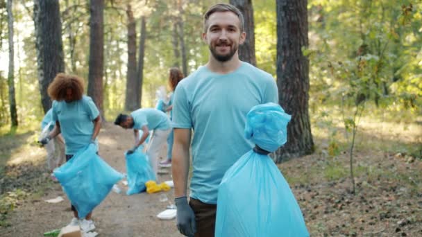 Miembro del equipo de voluntariado sosteniendo bolsa de basura con basura en el bosque sonriendo a la cámara — Vídeos de Stock