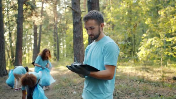 Член волонтерского движения занят бумажной работой в лесу, пока люди собирают мусор — стоковое видео