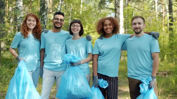 快乐的青年志愿者，背着垃圾袋站在森林里，微笑着拥抱 — 图库视频影像