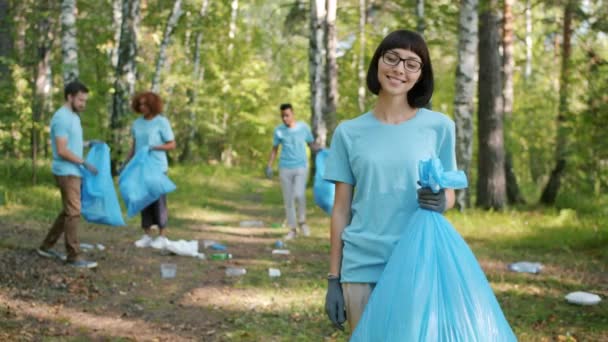 Söt kvinnlig volontär tittar på kameran håller papperskorgen i förorenad park — Stockvideo