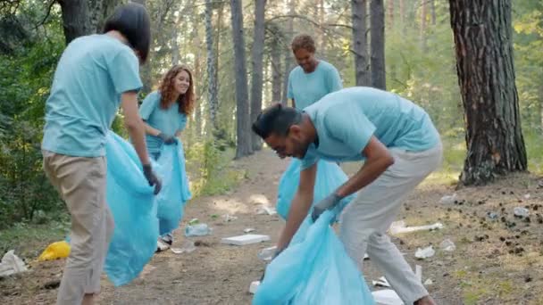 Śmiejący się młodzi ludzie zbierający śmieci w lesie sprzątający przyrodę bawiący się razem — Wideo stockowe