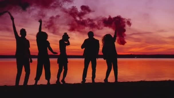 Les filles et les gars s'amusent à danser au coucher du soleil près de la rivière tenant des bombes fumigènes — Video