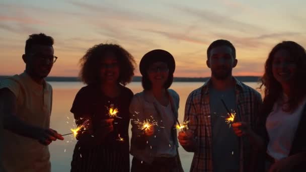Портрет счастливых людей, держащих бенгальские огни, стоящие на пляже вечером — стоковое видео