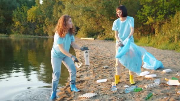 Весёлые девушки-волонтёры собирают мусор на пляже и болтают о уборке — стоковое видео