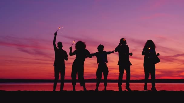 Siluetas de personas bailando con luces de bengala en la playa por la noche divirtiéndose — Vídeo de stock