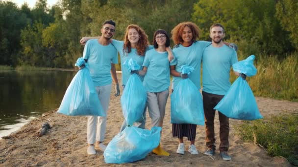 Önkéntesek többfajú csoportja szemeteszsákkal, akik mosolygva ölelkeznek a tisztított tavon. — Stock videók