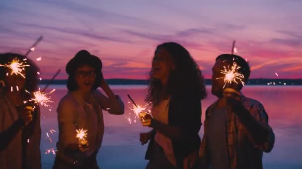 Smějící se přátelé tančí na jezeře beath s jiskry při západu slunce baví — Stock video