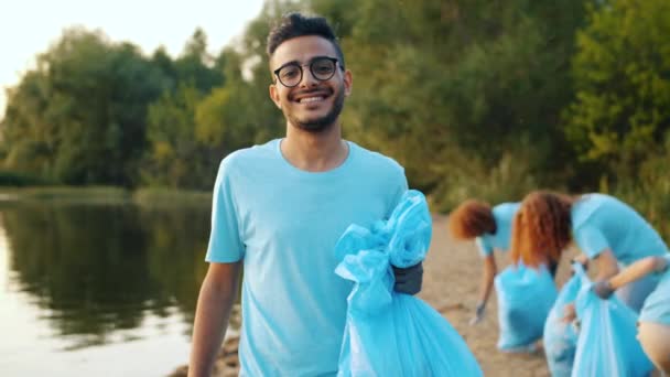 Fiatal arab önkéntes szemeteszsákkal mosolygott a tó partján, háttérben emberekkel. — Stock videók