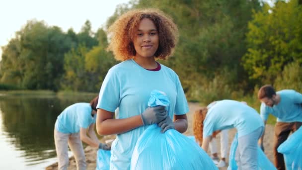 在被污染的湖岸提着垃圾袋微笑的非裔美国志愿者 — 图库视频影像