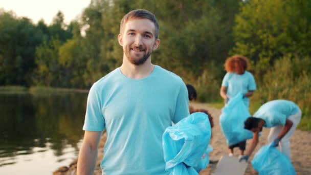 Szczęśliwy młody eko aktywista trzyma worek na śmieci patrząc na kamerę na zanieczyszczonej plaży jeziora — Wideo stockowe