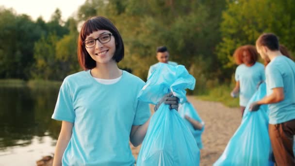 Radosna kobieta eko aktywistka trzyma worek na śmieci na brzegu jeziora z ciężko pracującym zespołem — Wideo stockowe