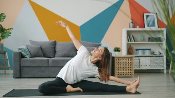 Bonita dama inclinada a una pierna estirando el cuerpo sentada en la esterilla de yoga en el apartamento — Vídeos de Stock