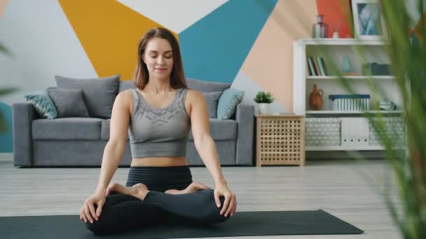 Женщина медитирует в позе лотоса сидя на коврике для йоги с закрытыми глазами дома — стоковое видео