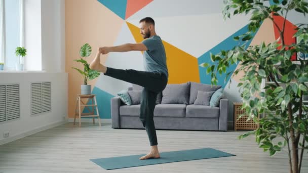 Männlicher Erwachsener übt Gleichgewichtsasana, steht auf einem Bein auf einer Matte und macht Yoga — Stockvideo