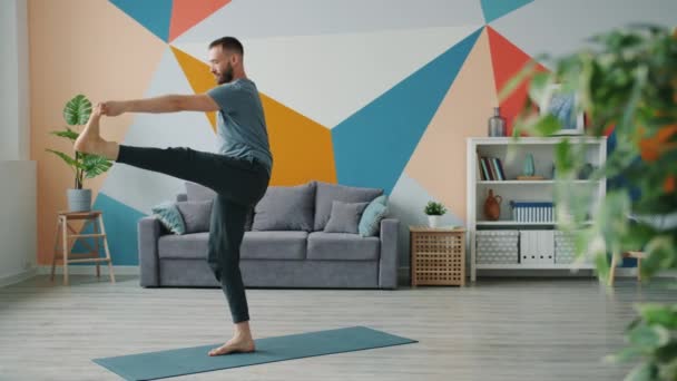 Медленное движение красивого мужчины, балансирующего на одной ноге, занимающегося йогой в квартире — стоковое видео