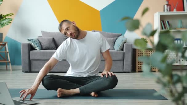 Hombre joven haciendo yoga en casa viendo tutoriales en línea en la pantalla del ordenador portátil — Vídeo de stock