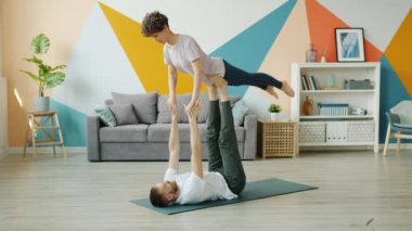 Mutlu çift evde yoga yapıyor, erkek yerde bacakları olan kadını tutuyor.
