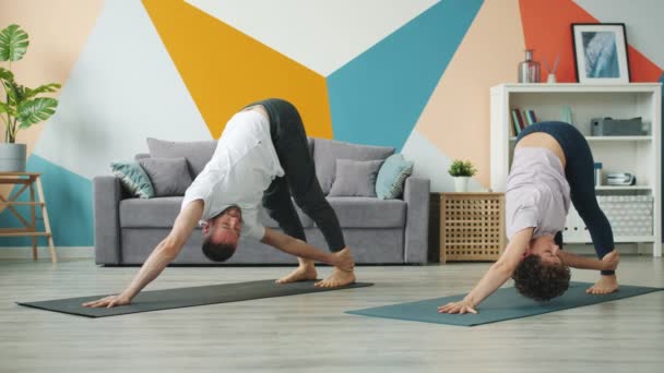 Girl and guy doing yoga in apartment together in downward dog position on mats — ストック動画