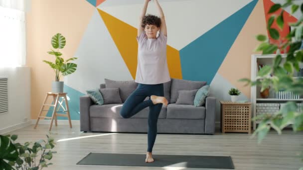 Bella dama disfrutando de la posición del árbol haciendo yoga en casa de pie en la habitación en la estera — Vídeos de Stock