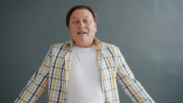 Грайливий азіатський чоловік посміхається, коли веселиться на сірому фоні. — стокове відео