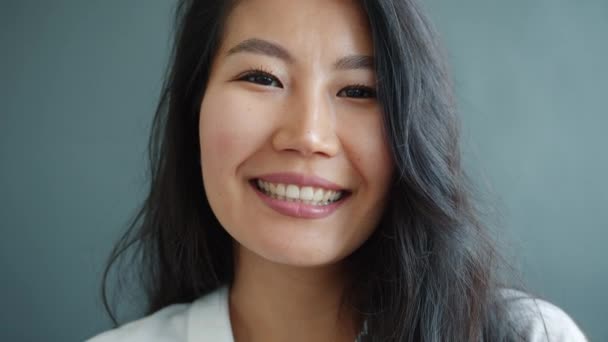Közelkép egy csinos ázsiai hölgyről, aki szürke háttéren mosolyog. — Stock videók