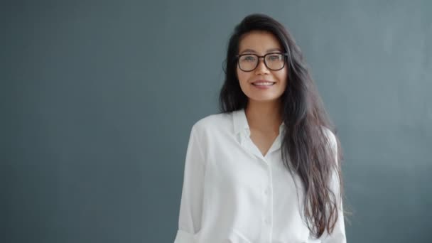 Retrato de mujer asiática feliz agitando la mano sonriendo en el interior sobre fondo gris — Vídeos de Stock