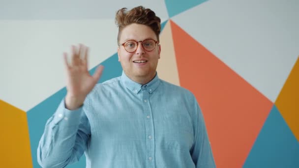 Смішний молодий чоловік в окулярах махає рукою з радісним обличчям на барвистому фоні — стокове відео