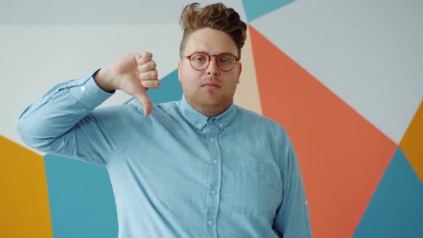 Unhappy stylish guy showing thumbs-down gesture smiling in funny way indoors — 비디오